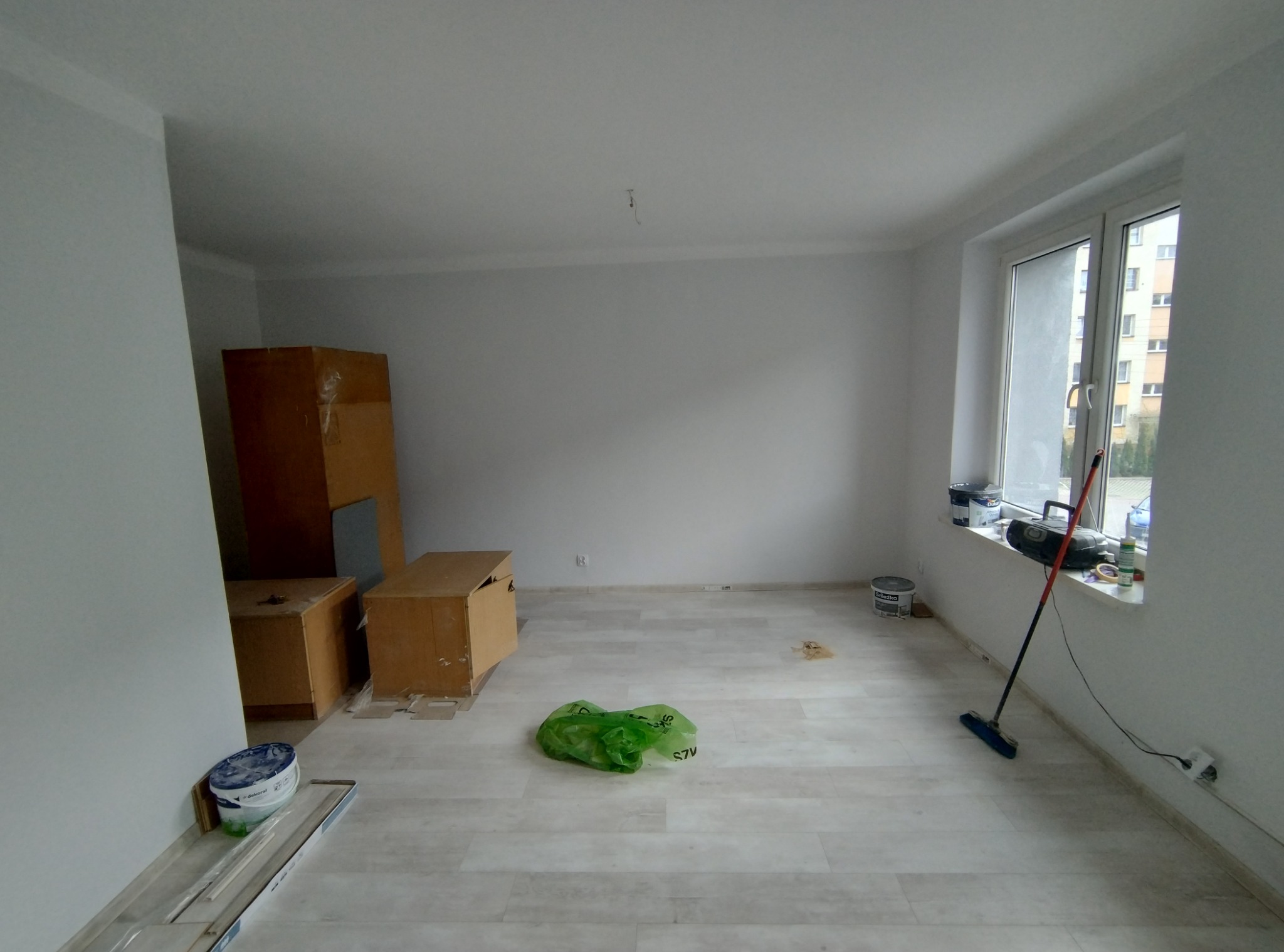 Pomalowane na biało puste pomieszczenie z panelami podłogowymi, skrzyniami kartonowymi, wiadrem z farbą przy ścianie, workiem na śmieci na podłodze i mopem opartym o ścianę przy oknie.