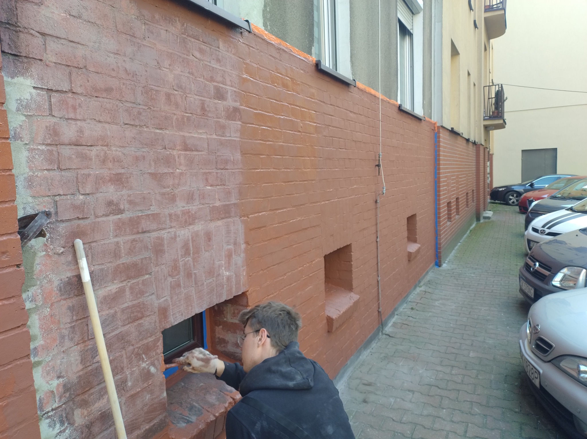 Malowanie ceglanej ściany zewnętrznej w trakcie realizacji; widoczna częściowo pomalowana powierzchnia, mężczyzna w trakcie malowania okna, zabezpieczonego niebieską taśmą malarską.
