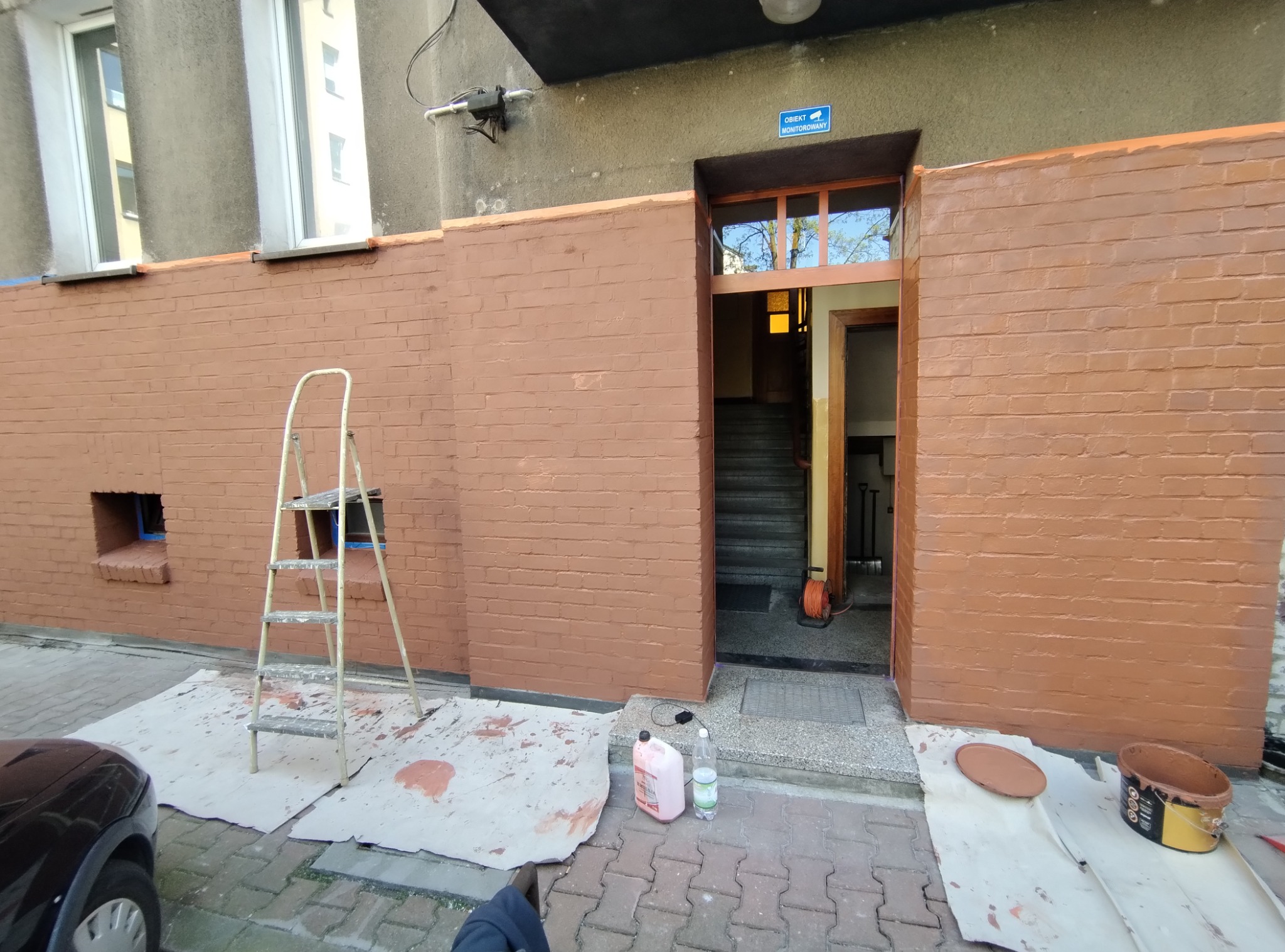 Malowanie elewacji budynku z cegły na brązowo, widoczna drabina, pojemniki z farbą i ślady farby na podłożu.