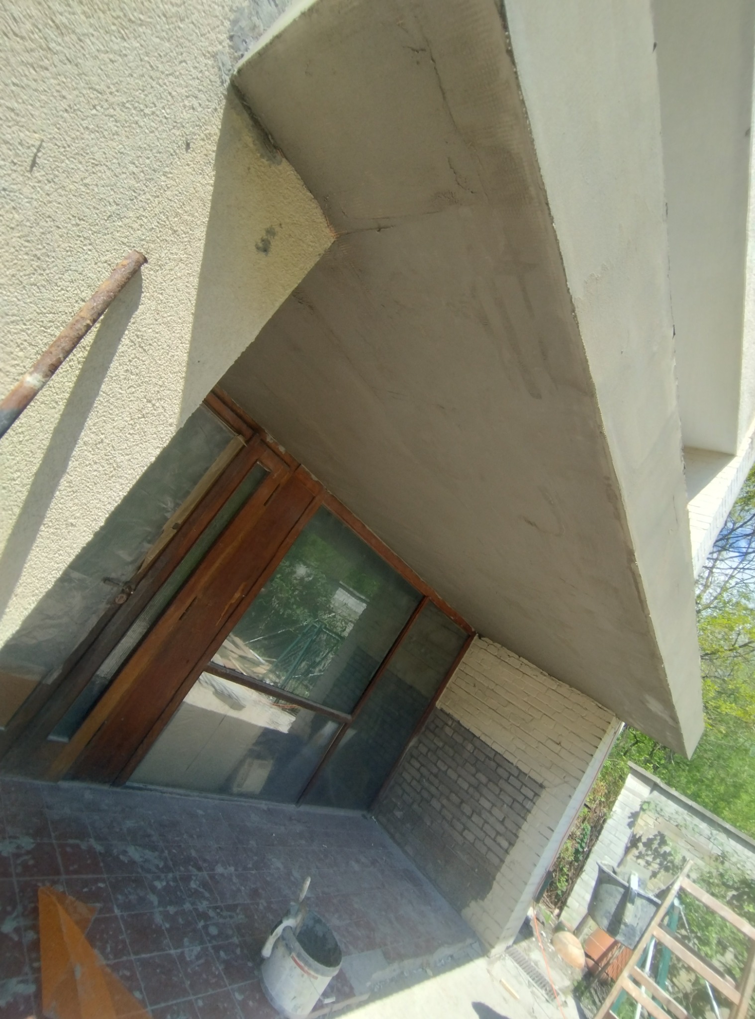 Zewnętrzny remont balkonu z drewnianą stolarką okienną, widoczna ściana z cegły pomalowana do połowy, wiadro i narzędzia malarskie na kafelkach.