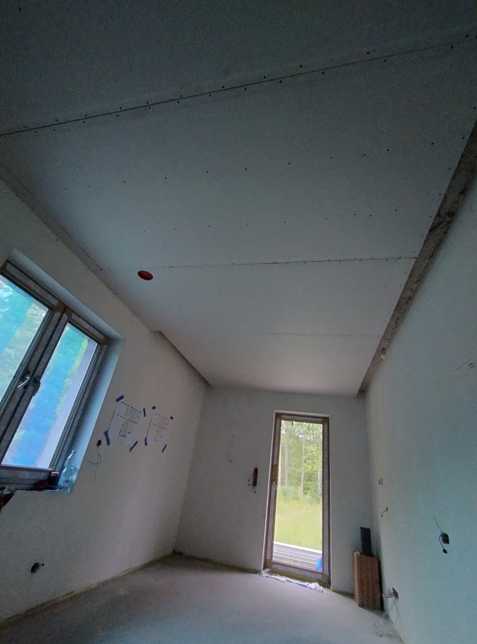 Wnętrze pokoju w trakcie remontu: zamontowane płyty karton gips na suficie, widoczne mocowania, na ścianie przyklejone plany mieszkania, okno dachowe z widokiem na zieleń.