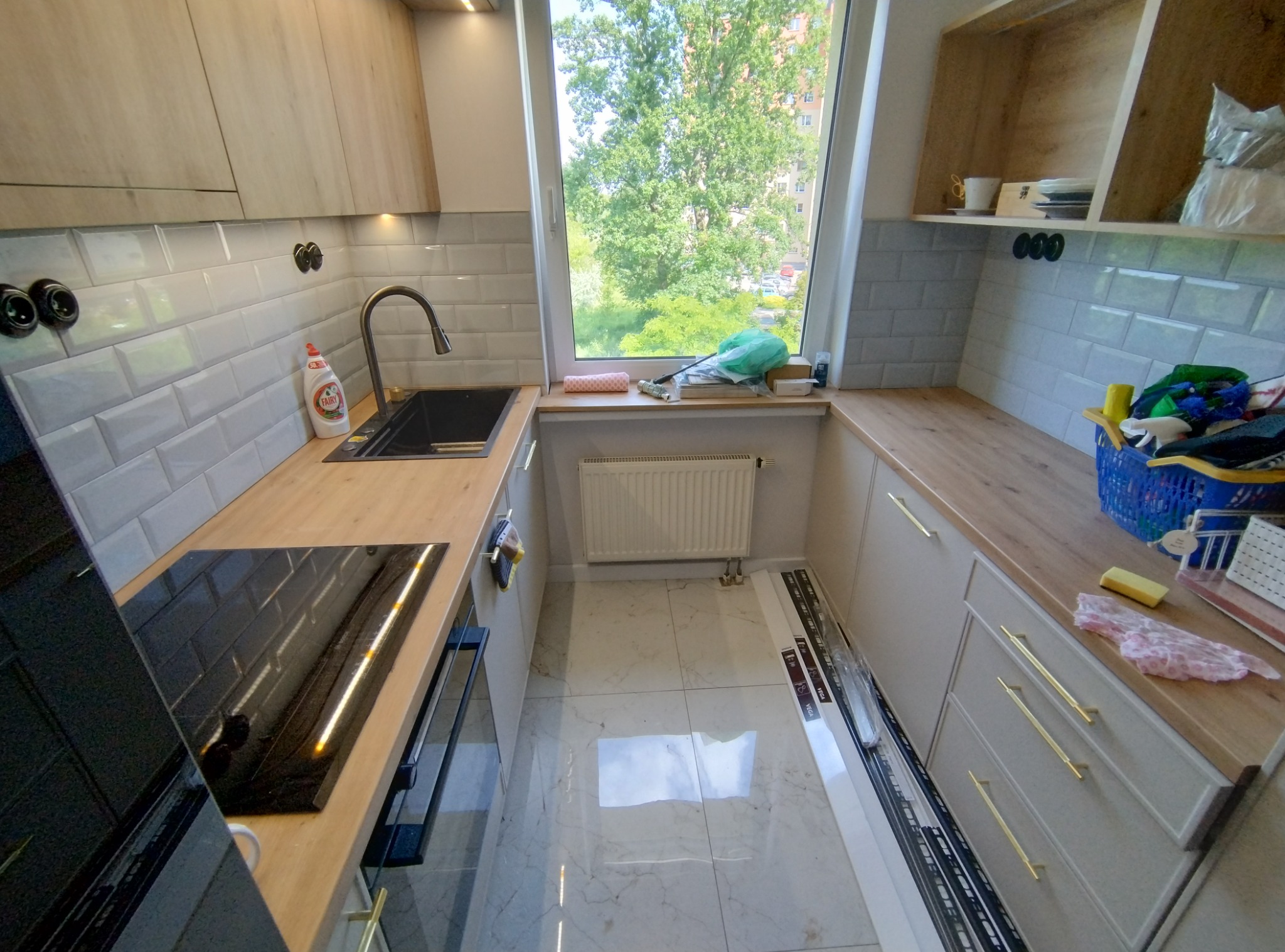 Nowoczesna kuchnia z jasnymi drewnianymi szafkami, szarymi płytkami metra, czarnym zlewem i płytą indukcyjną, widok z okna na zieleń drzew.