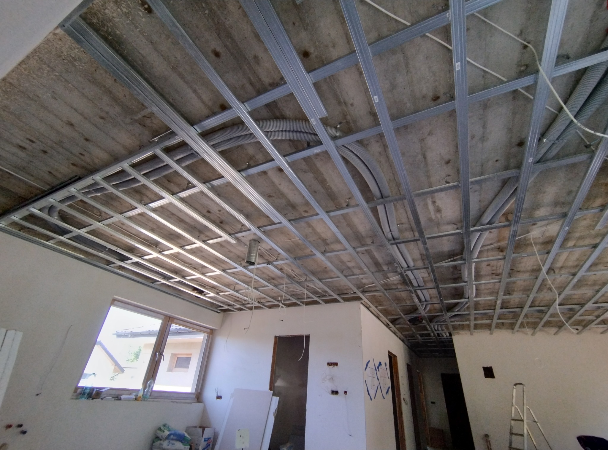 Metalowa konstrukcja sufitu podwieszanego z widocznymi rurami wentylacyjnymi i przewodami elektrycznymi, surowy stan przed montażem płyt gipsowo-kartonowych, widok z dołu.