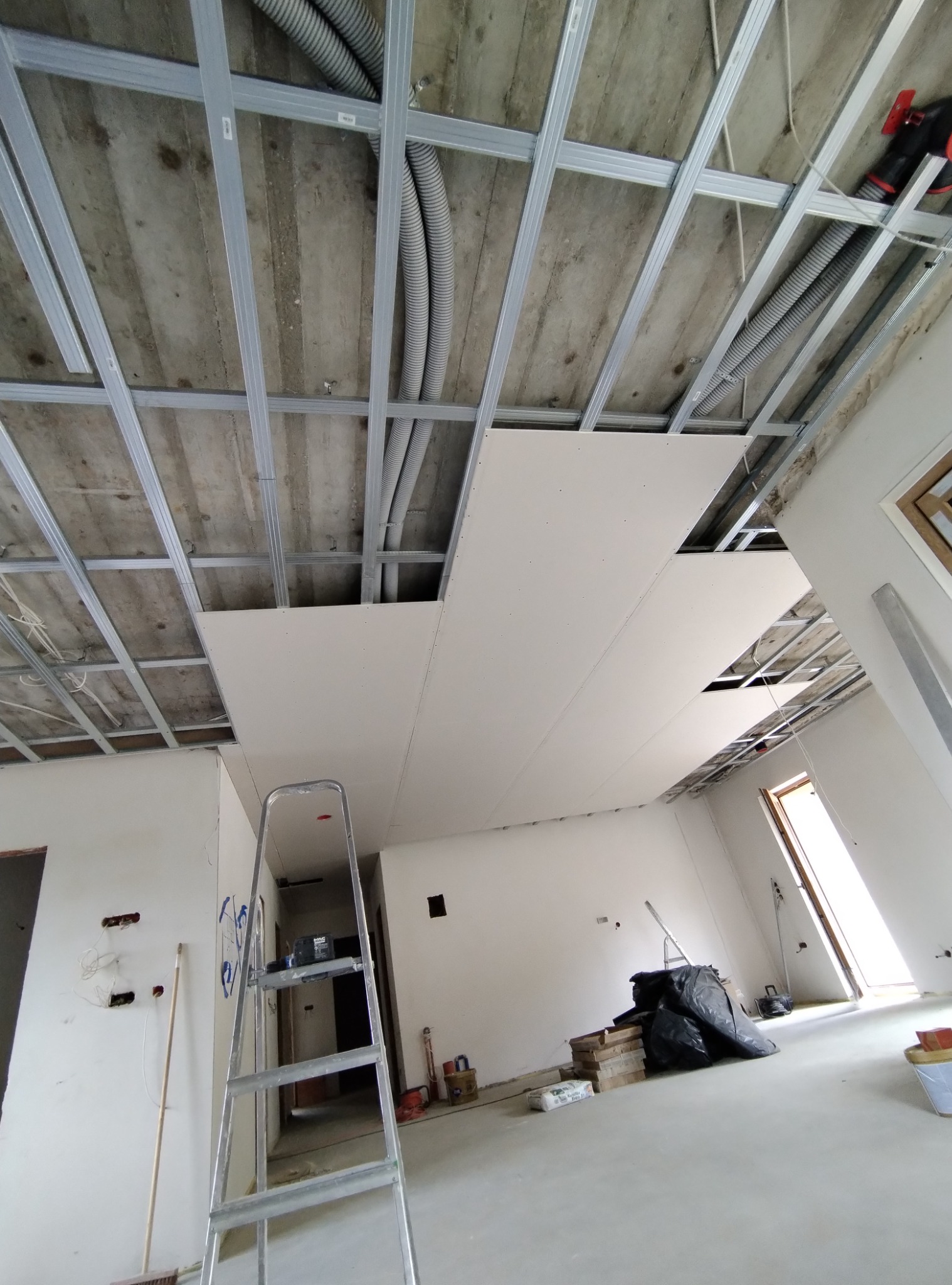 Widok z dołu na częściowo zamontowany sufit podwieszany z płyt kartonowo-gipsowych na metalowej konstrukcji, widoczne kable w peszlach, drabina aluminiowa w pomieszczeniu w stanie surowym.