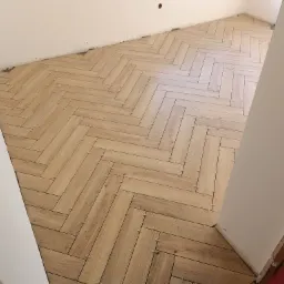 Układanie podłogi z płytek imitujących drewno w jodełkę, widoczne krzyżyki dystansowe, surowy stan ścian.