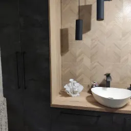 Nowoczesna łazienka z umywalką nablatową, armaturą w kolorze czarnym, drewnopodobnymi płytkami ułożonymi w jodełkę na ścianie oraz szafkami w kolorze grafitowym.