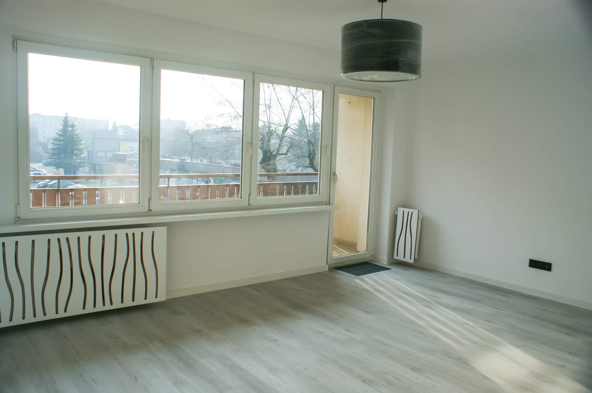Pusty pokój po remoncie z panelami podłogowymi, grzejnikiem z dekoracyjną obudową, oknami z widokiem na balkon i miasto oraz lampą wiszącą.