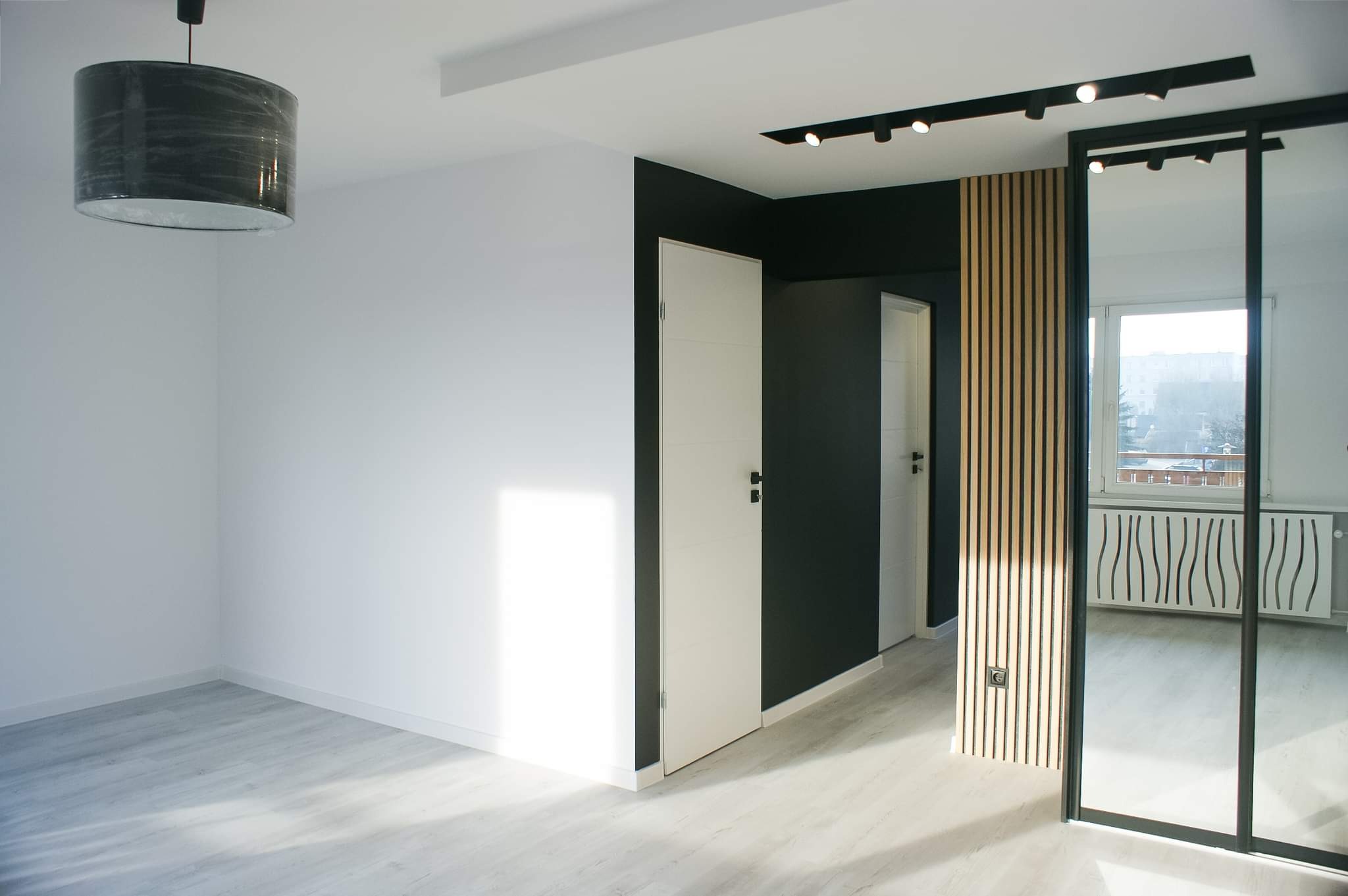 Nowoczesne wnętrze po remoncie: białe ściany, szare panele podłogowe, czarna ściana z białymi drzwiami, drewniana lamelowa ściana dekoracyjna z gniazdkiem, czarne oświetlenie sufitowe i lampa...