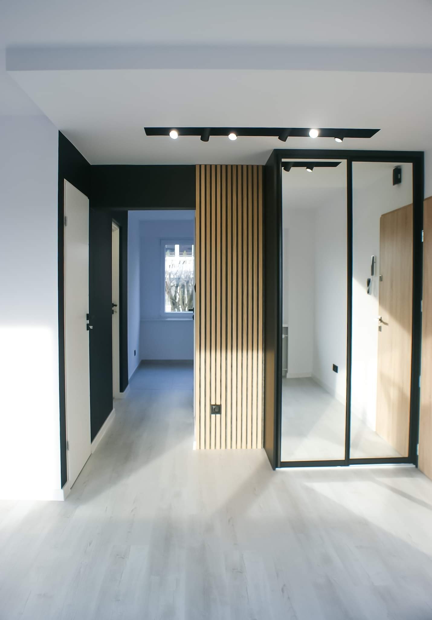 Nowoczesne wnętrze mieszkania z drewnianą ścianką działową z lameli, czarną listwą sufitową z reflektorami punktowymi, szklanymi drzwiami z czarną ramą i jasną podłogą.