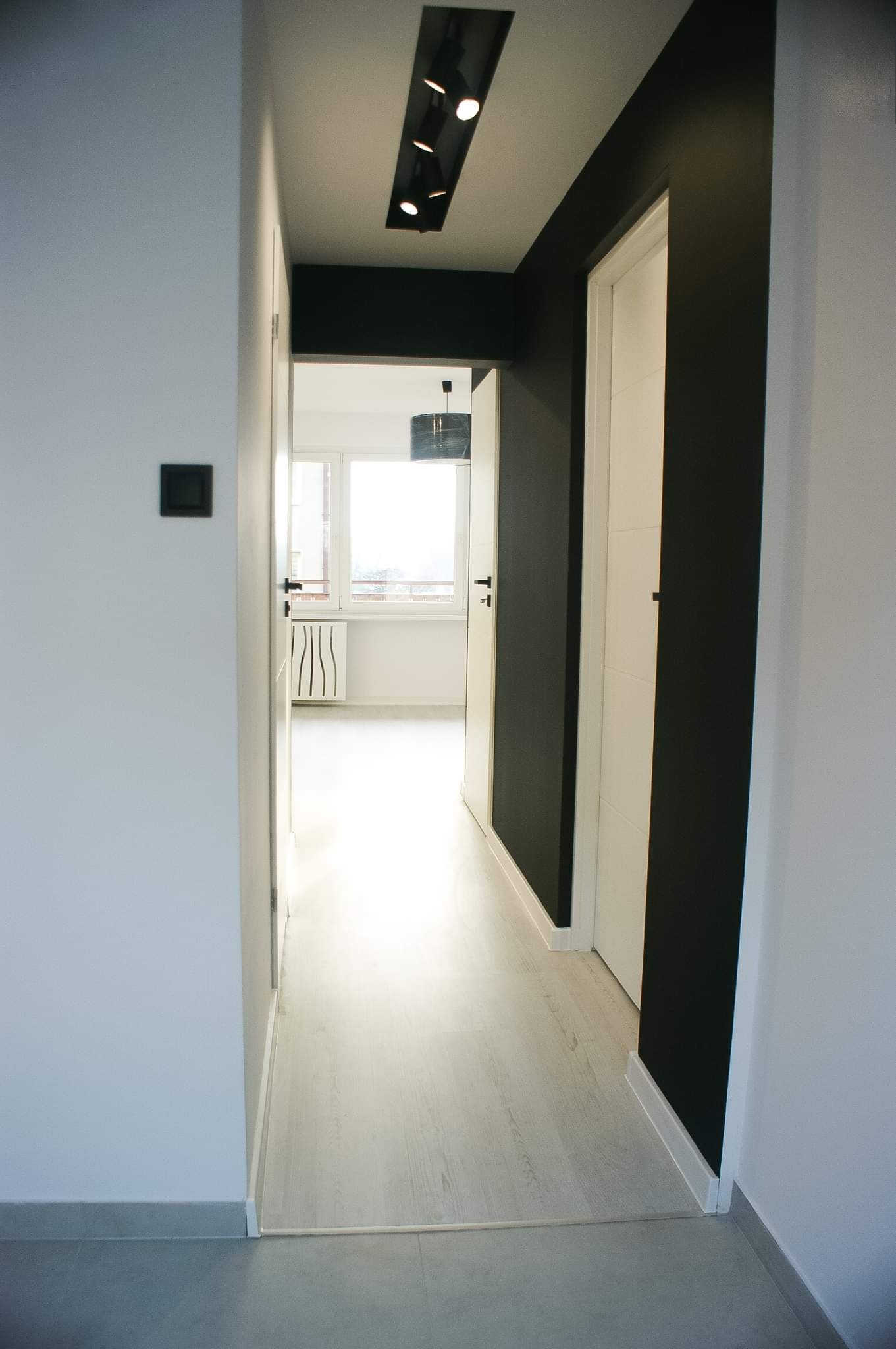 Nowoczesny korytarz z białymi ścianami i panelami podłogowymi, kontrastująca czarna ściana z białymi drzwiami, oświetlenie szynowe sufitowe i widok na jasny pokój z grzejnikiem.