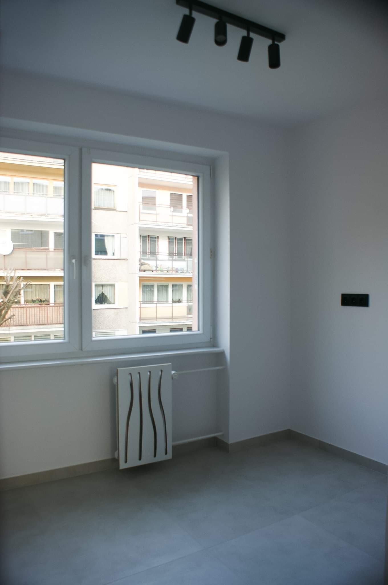 Pusty, odnowiony pokój z szarymi płytkami podłogowymi, białymi ścianami, nowoczesnym grzejnikiem dekoracyjnym, oknem z widokiem na budynek mieszkalny i czarną lampą sufitową