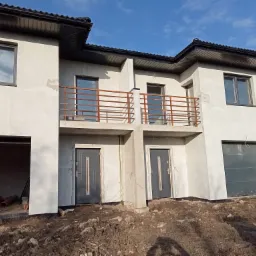 Dwukondygnacyjny dom w stanie surowym otwartym z balkonami i garażem, widoczne worki z zaprawą i rury na ziemi.