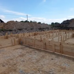 Szalunek z surowego drewna przygotowany do wylania fundamentów domu, widoczne wykopane podłoże i pryzma ziemi, w tle fragment budynku mieszkalnego pod błękitnym niebem.