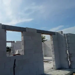 Wznoszenie ścian domu z bloczków betonu komórkowego Ytong z widocznymi nadprożami okiennymi i zbrojeniami pionowymi, na tle błękitnego nieba.