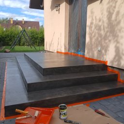 Liubomyr Ovchar - Zewnętrzne schody wejściowe wykończone szarym, cieniowanym mikrocementem, zabezpieczone pomarańczową taśmą malarską, obok rozłożone narzędzia i puszki z preparatami na kartonie, w tle dom...