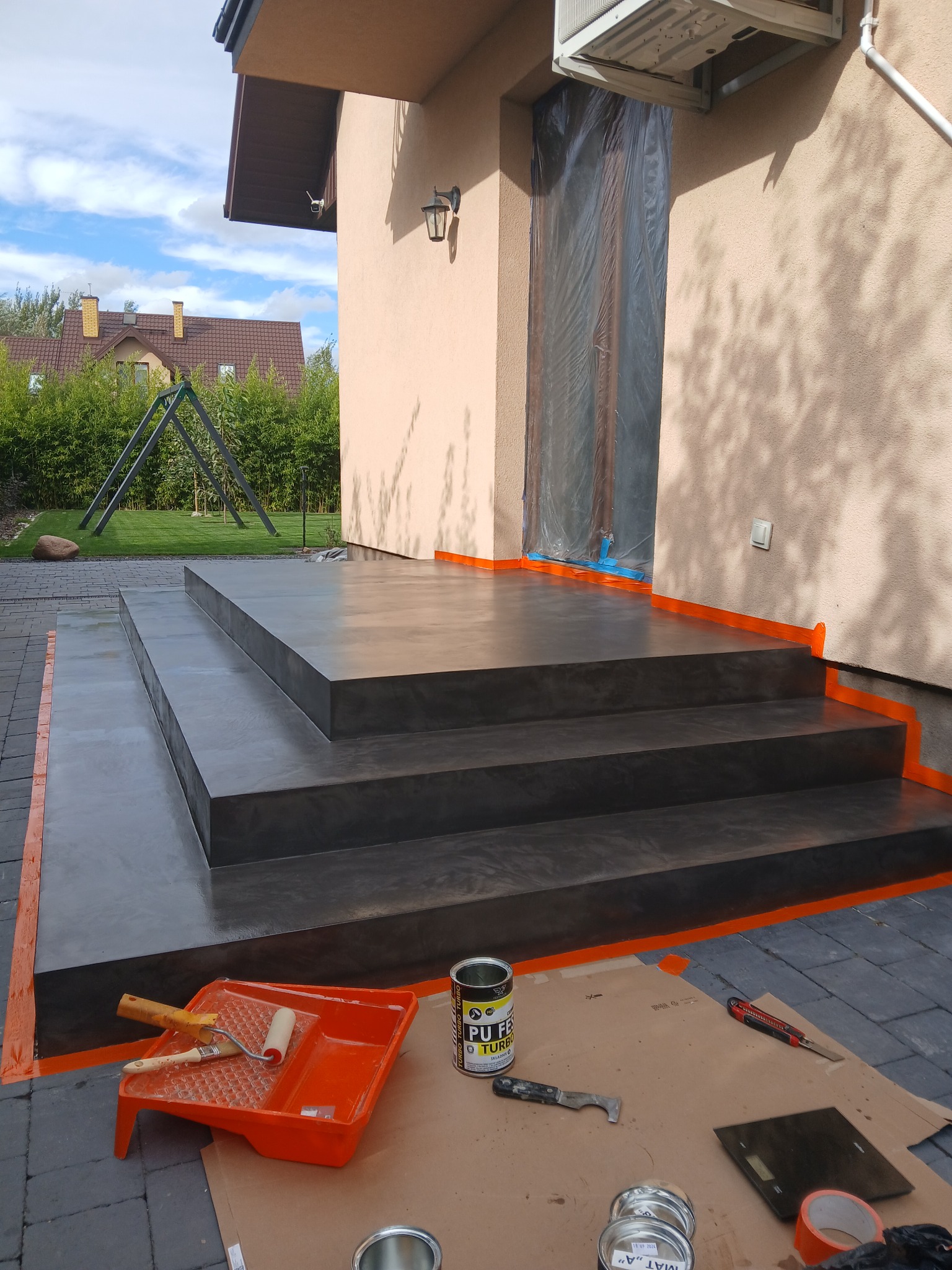 Zewnętrzne schody wejściowe wykończone szarym, cieniowanym mikrocementem, zabezpieczone pomarańczową taśmą malarską, obok rozłożone narzędzia i puszki z preparatami na kartonie, w tle dom...