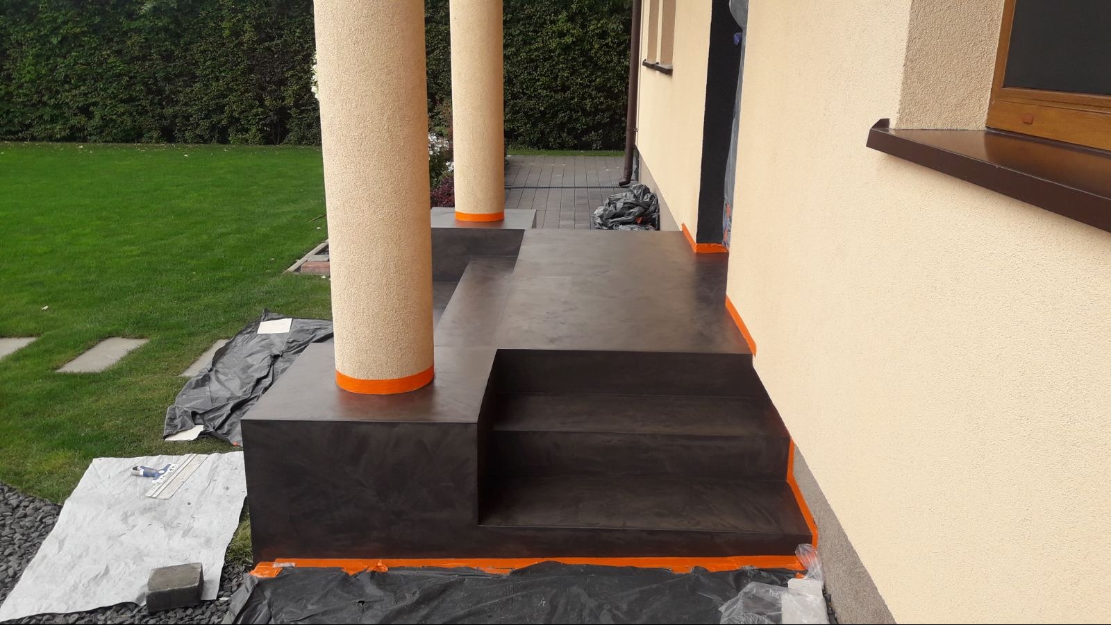 Zewnętrzne schody wejściowe i taras wykończone ciemnym, polerowanym materiałem z pomarańczowymi akcentami, widoczne kolumny z fakturą i zabezpieczenia folią malarską.