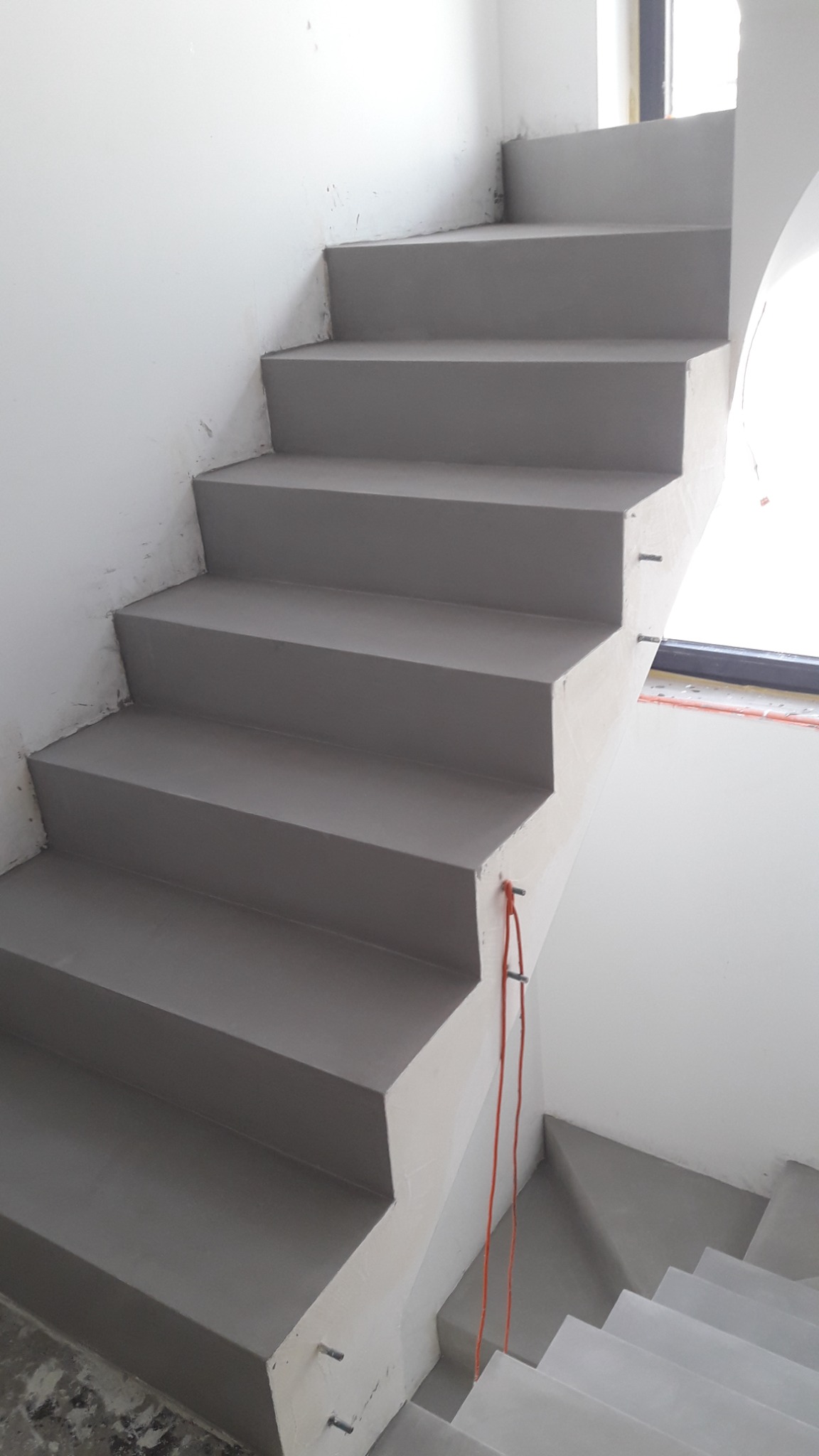 Schody betonowe w szarym odcieniu, świeżo wykończone, z widocznymi metalowymi elementami mocującymi i pomarańczowym kablem zwisającym wzdłuż ściany, w surowym stanie.