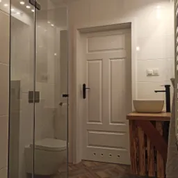 Nowoczesna łazienka z kabiną prysznicową ze szklanymi drzwiami, białą ceramiką, drewnianą szafką pod umywalką i wzorzystą podłogą z płytek.