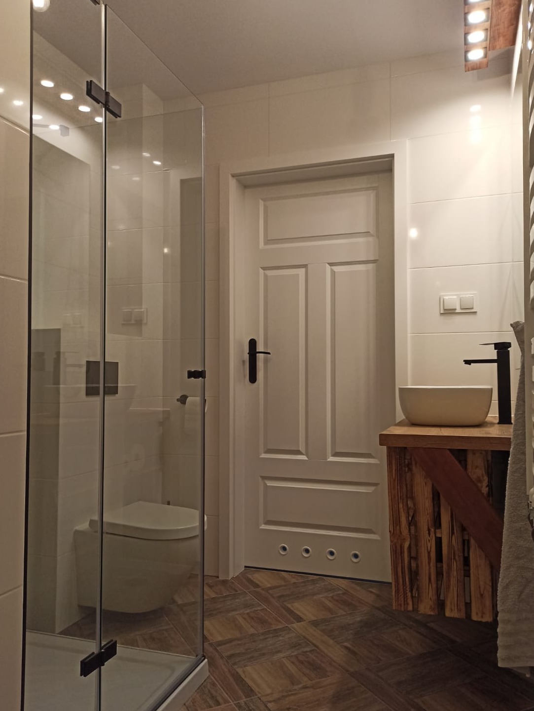 Nowoczesna łazienka z kabiną prysznicową ze szklanymi drzwiami, białą ceramiką, drewnianą szafką pod umywalką i wzorzystą podłogą z płytek.