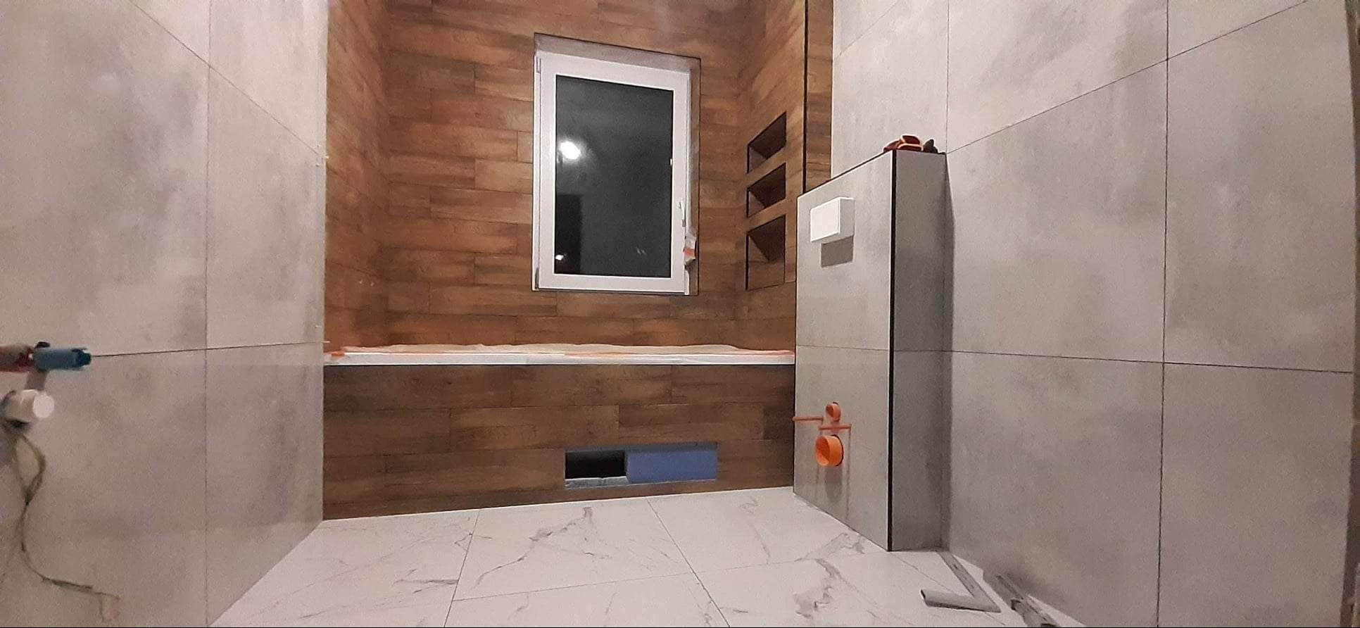 Wnętrze łazienki w trakcie prac wykończeniowych: ściany częściowo pokryte szarymi płytkami wielkoformatowymi, fragment ściany wykończony panelami drewnopodobnymi, widoczne instalacje hydrauliczne...