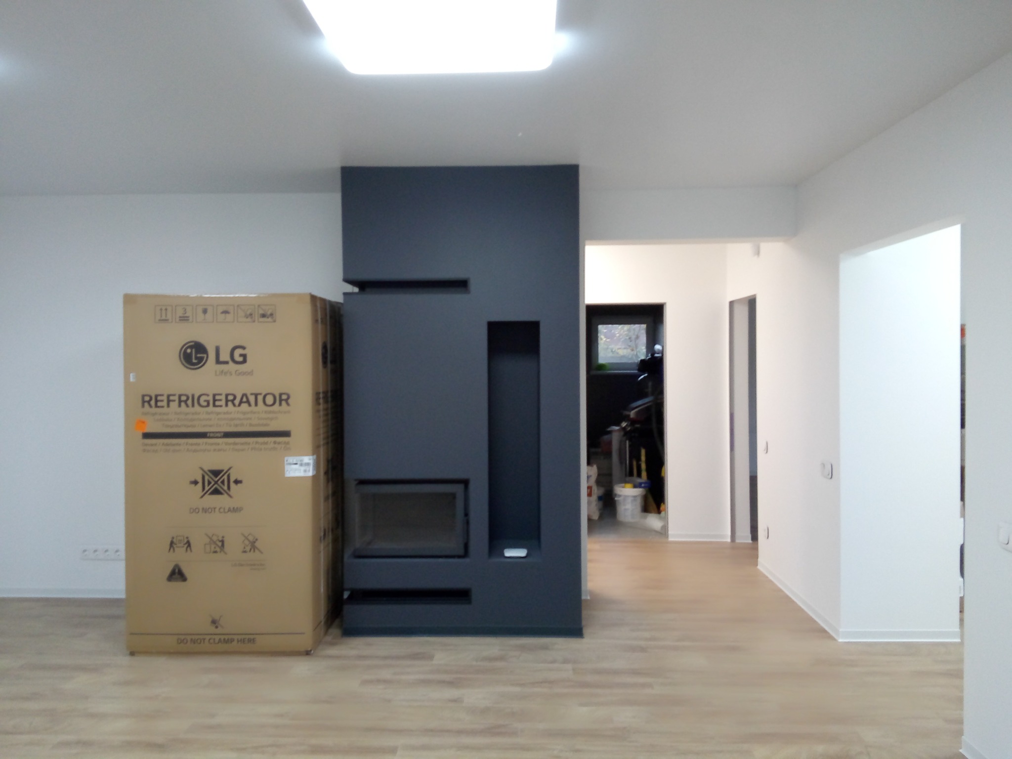 Nowoczesny, szary kominek z wkładem, ustawiony w minimalistycznym wnętrzu z jasnymi ścianami i drewnopodobną podłogą, obok stoi kartonowe opakowanie lodówki LG, widok na dalsze pomieszczenia.