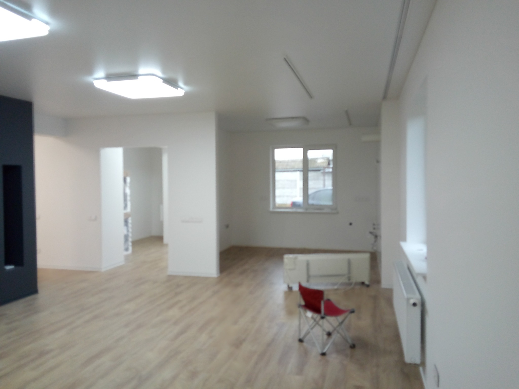 Jasne, przestronne wnętrze po remoncie z panelami podłogowymi, białymi ścianami i grzejnikami, oświetlone lampami sufitowymi; w tle widoczne okno i czerwone krzesło turystyczne.