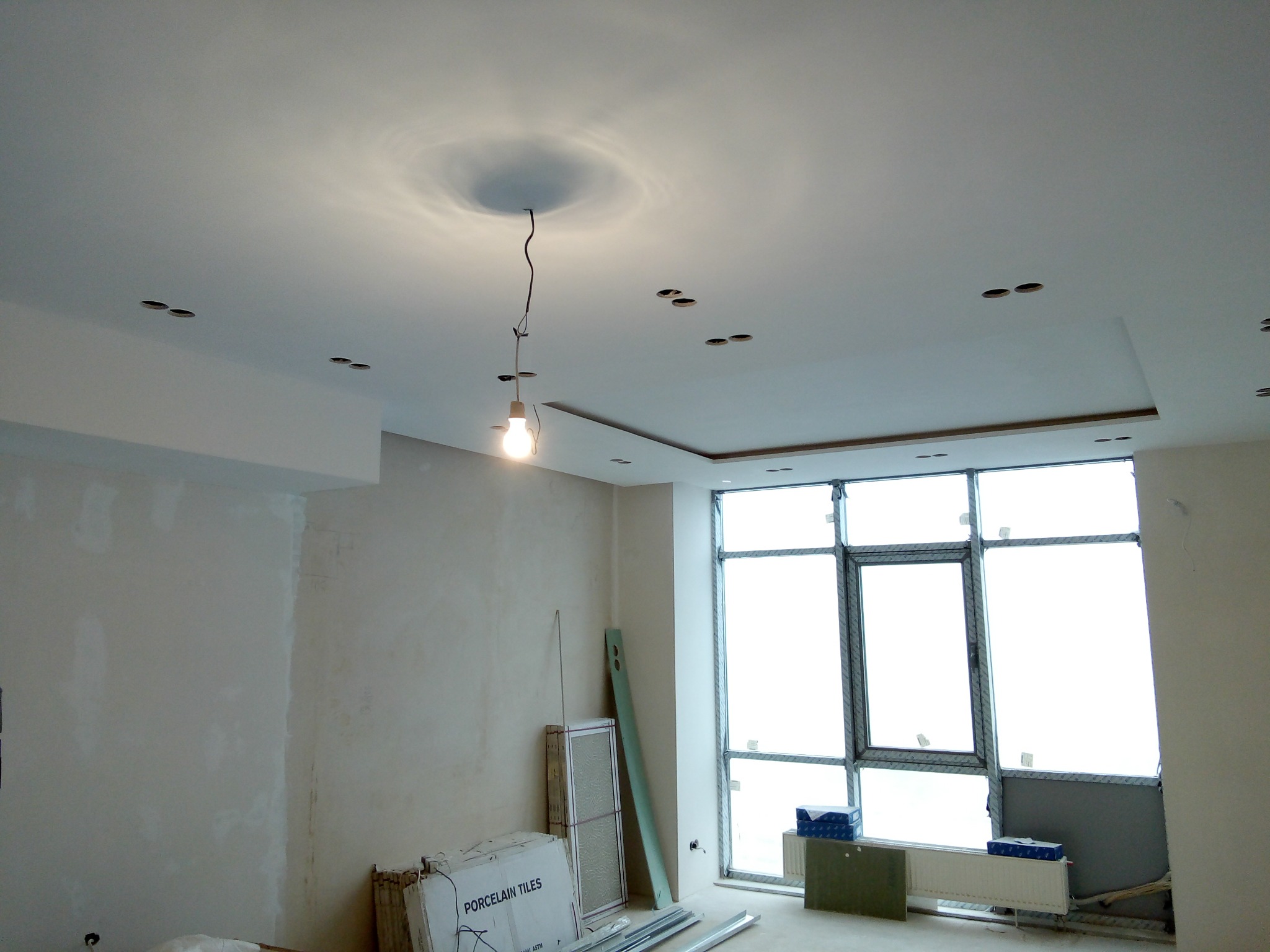 Wnętrze w trakcie prac wykończeniowych: surowa ściana, przygotowane otwory na oświetlenie w suficie podwieszanym, duże okno z widokiem na zewnątrz, paczki z płytkami ceramicznymi przy ścianie.