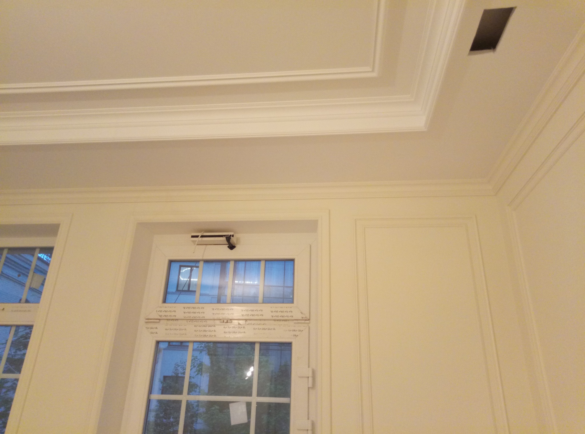 Jasne wnętrze z białymi ścianami, sztukaterią na suficie i oknami z widokiem na zewnątrz, podkreślone subtelnym oświetleniem.