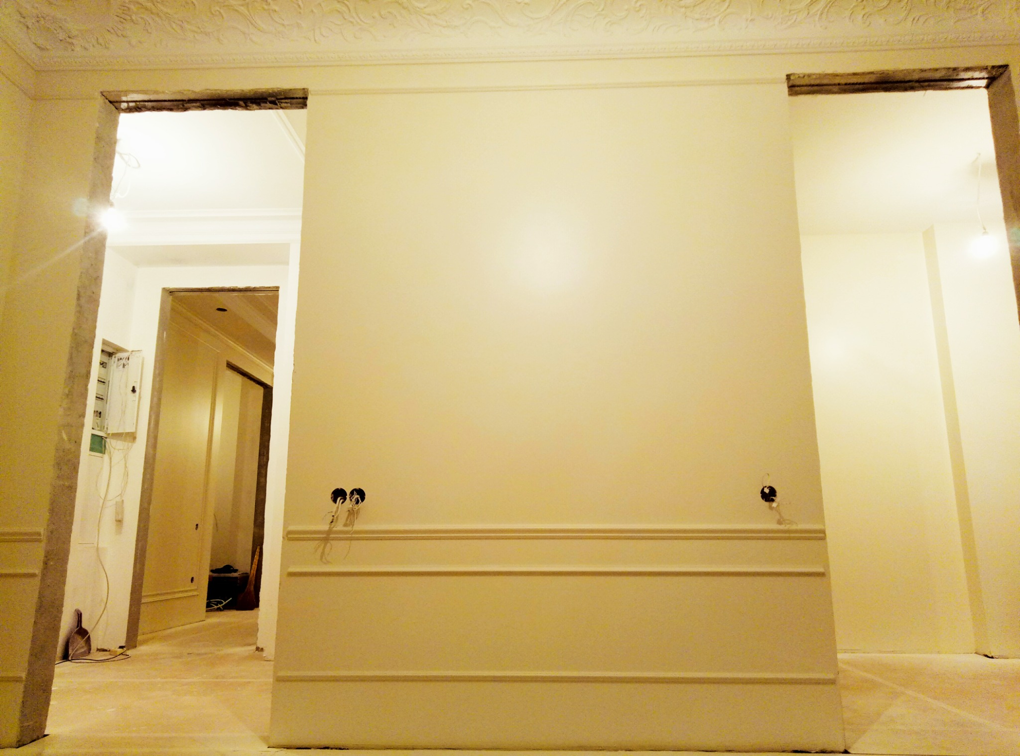 Ściana w trakcie prac wykończeniowych, widoczne listwy dekoracyjne, przygotowane punkty elektryczne i surowa podłoga, zarysowane przejścia do innych pomieszczeń.