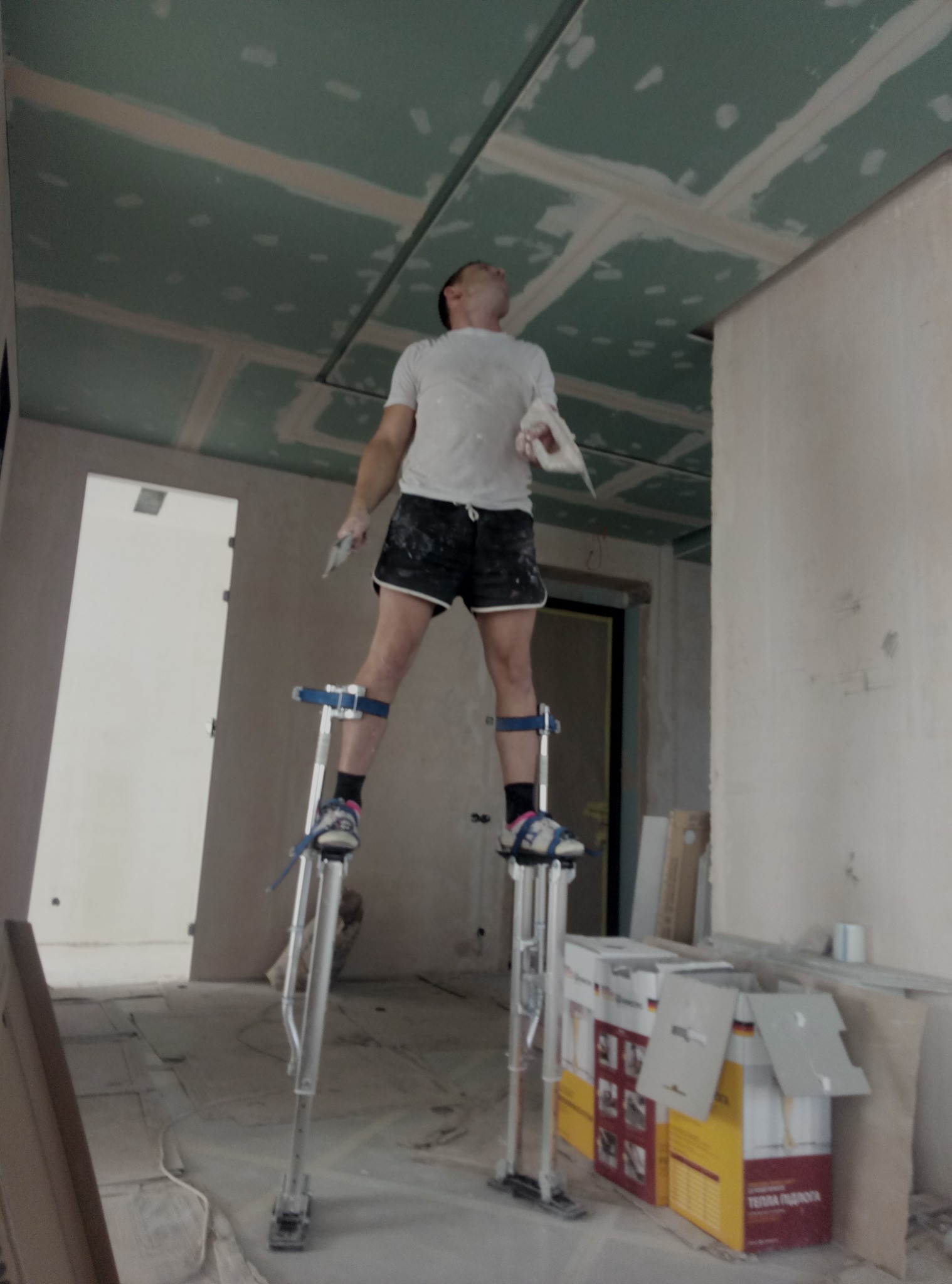 Pracownik na szczudłach szpachluje sufit z płyt gipsowo-kartonowych, widoczne łączenia płyt przed wygładzeniem, kartony z materiałami budowlanymi w tle.