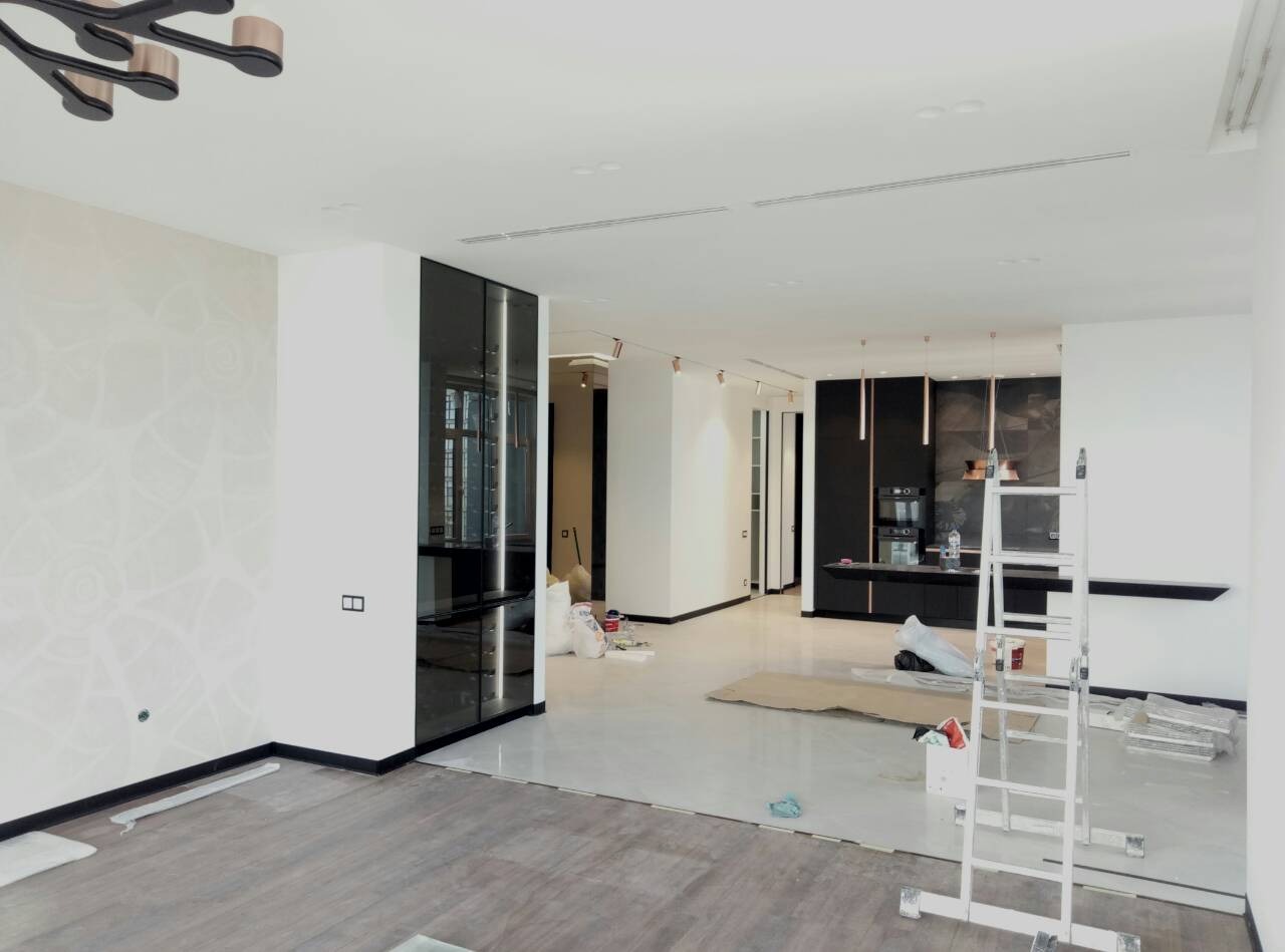 Wnętrze w trakcie prac wykończeniowych: częściowo położona podłoga z paneli, ściany pomalowane na biało z dekoracyjnym wzorem, widoczna drabina, zabezpieczona folią podłoga, worki z materiałami...