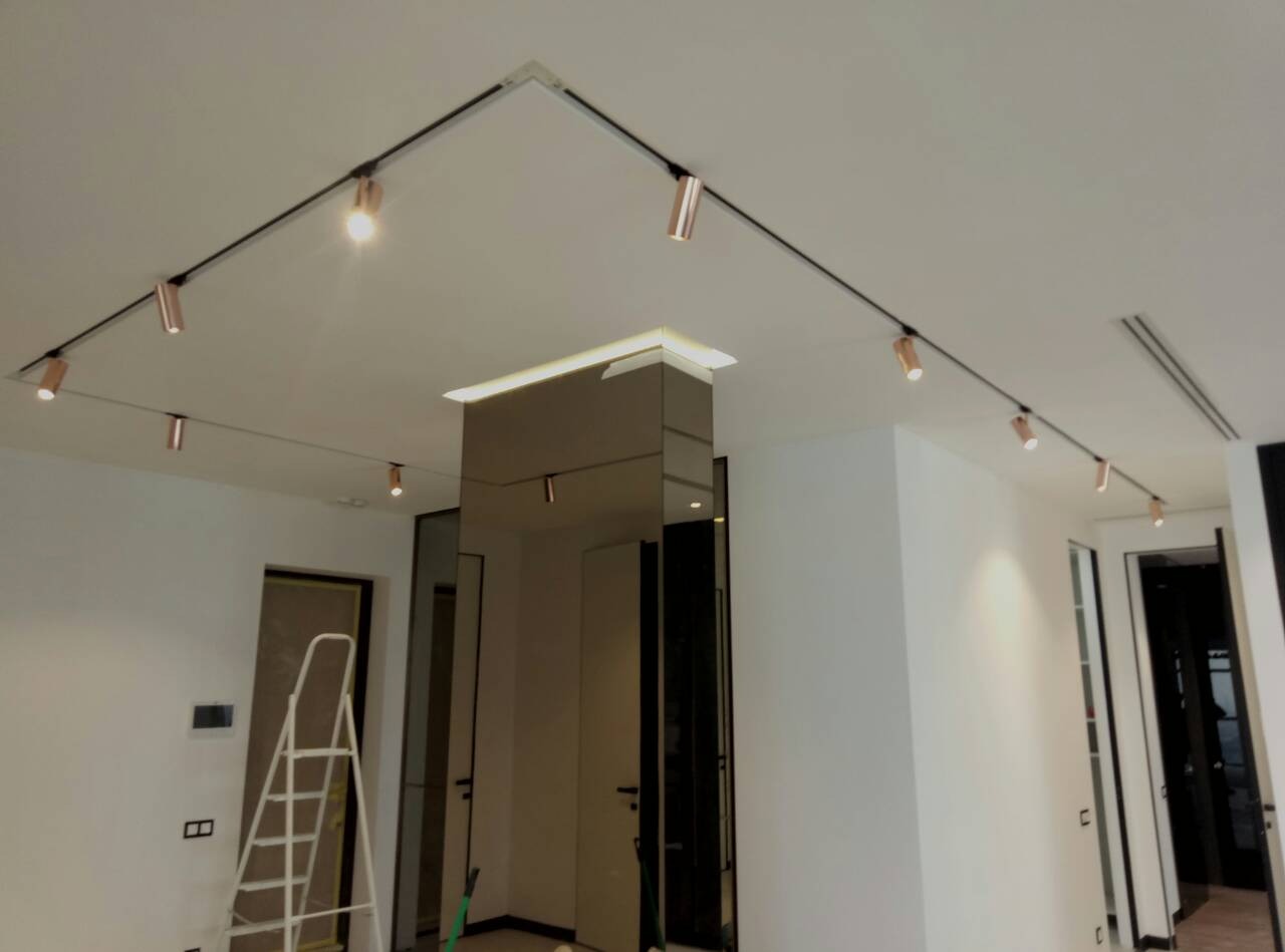 Nowoczesne oświetlenie szynowe z reflektorami w kolorze miedzi zamontowane na suficie w minimalistycznym wnętrzu, widoczna drabina malarska w tle sugerująca trwające prace wykończeniowe.