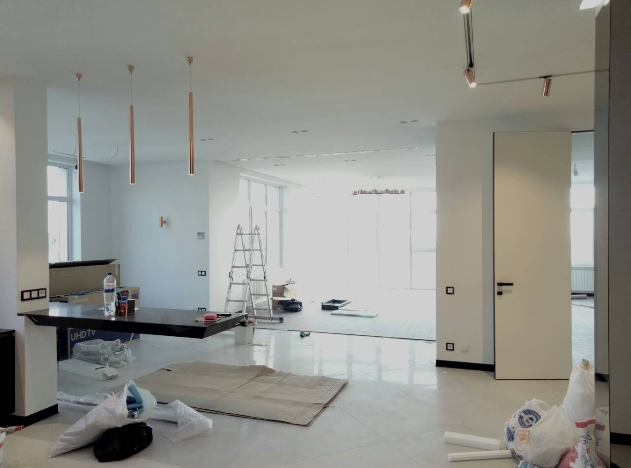 Przestronne wnętrze w trakcie remontu: białe ściany, marmurowe płytki podłogowe, nowoczesne lampy wiszące, rozłożone materiały budowlane i drabina w tle, wskazujące na trwające prace wykończeniowe.