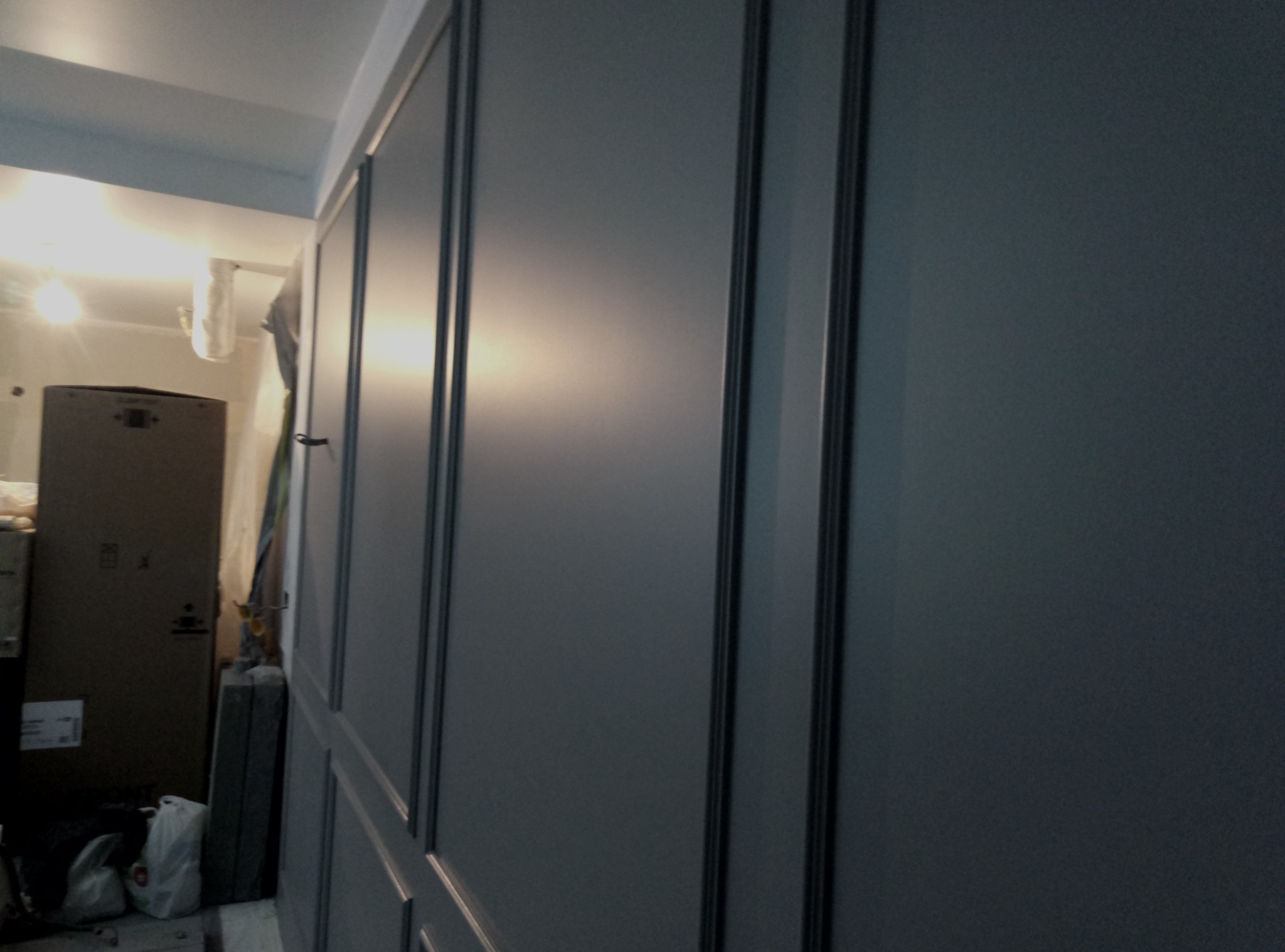 Panelowa ściana w odcieniach szarości z widocznymi pionowymi listwami, w tle kartonowe pudła i torby, prawdopodobnie w trakcie prac wykończeniowych.