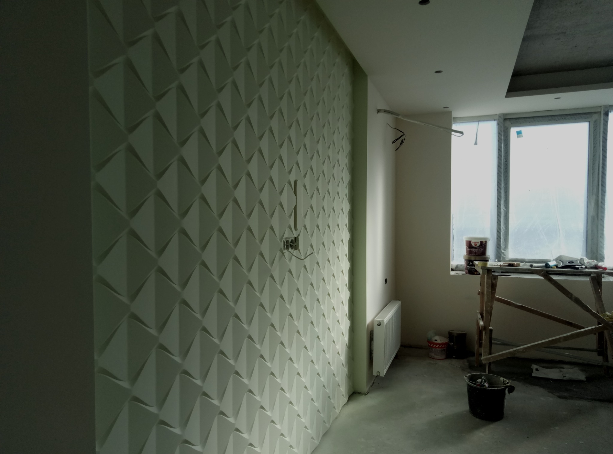 Ściana pokryta białą, trójwymiarową tapetą geometryczną w kształcie piramidek; widoczny fragment grzejnika, okno z folią ochronną i rusztowanie malarskie.