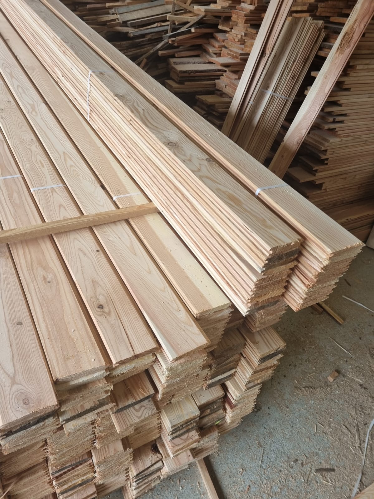 Stosy świeżo obrobionej, jasnej tarcicy ułożone w magazynie, widoczne naturalne słoje drewna i drobne wióry.