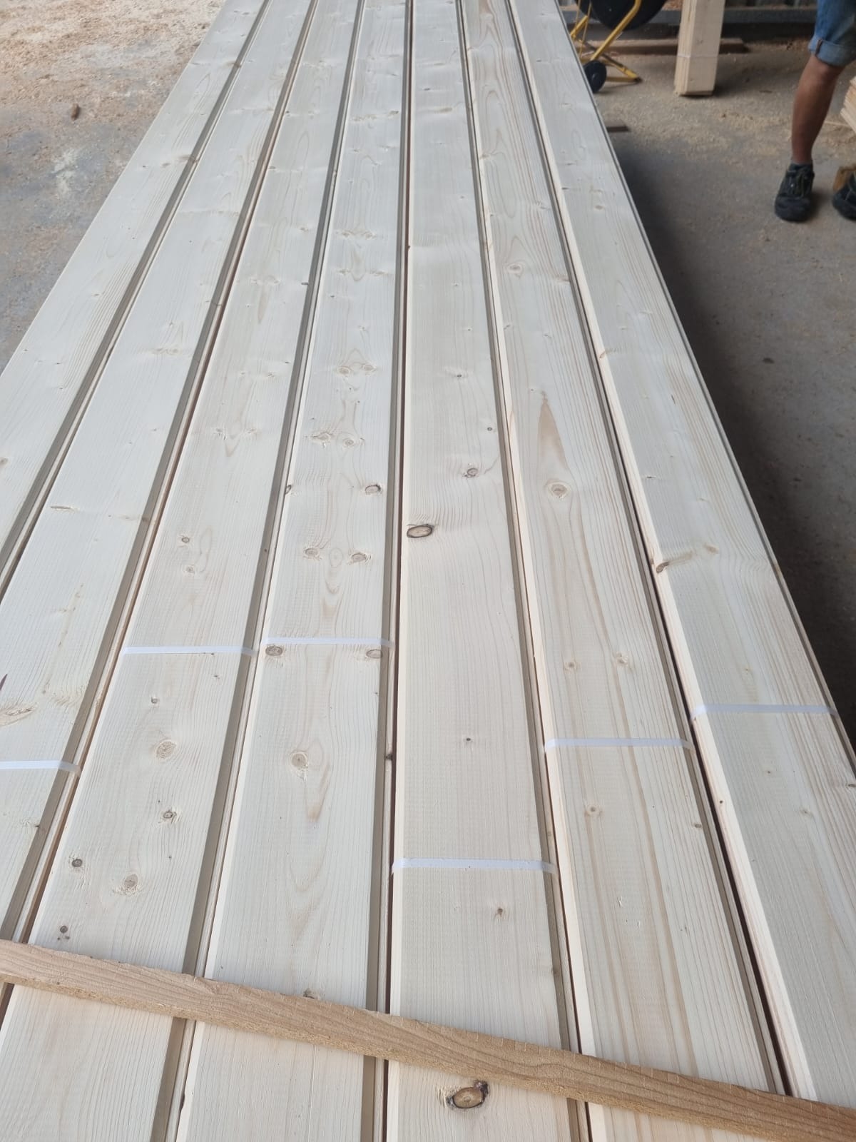 Stos jasnych, świeżo przetartych desek z widocznymi sękami, zabezpieczonych taśmą, ułożonych na szarym betonowym podłożu, z fragmentem drewnianej listwy na dole i fragmentem osoby w tle.