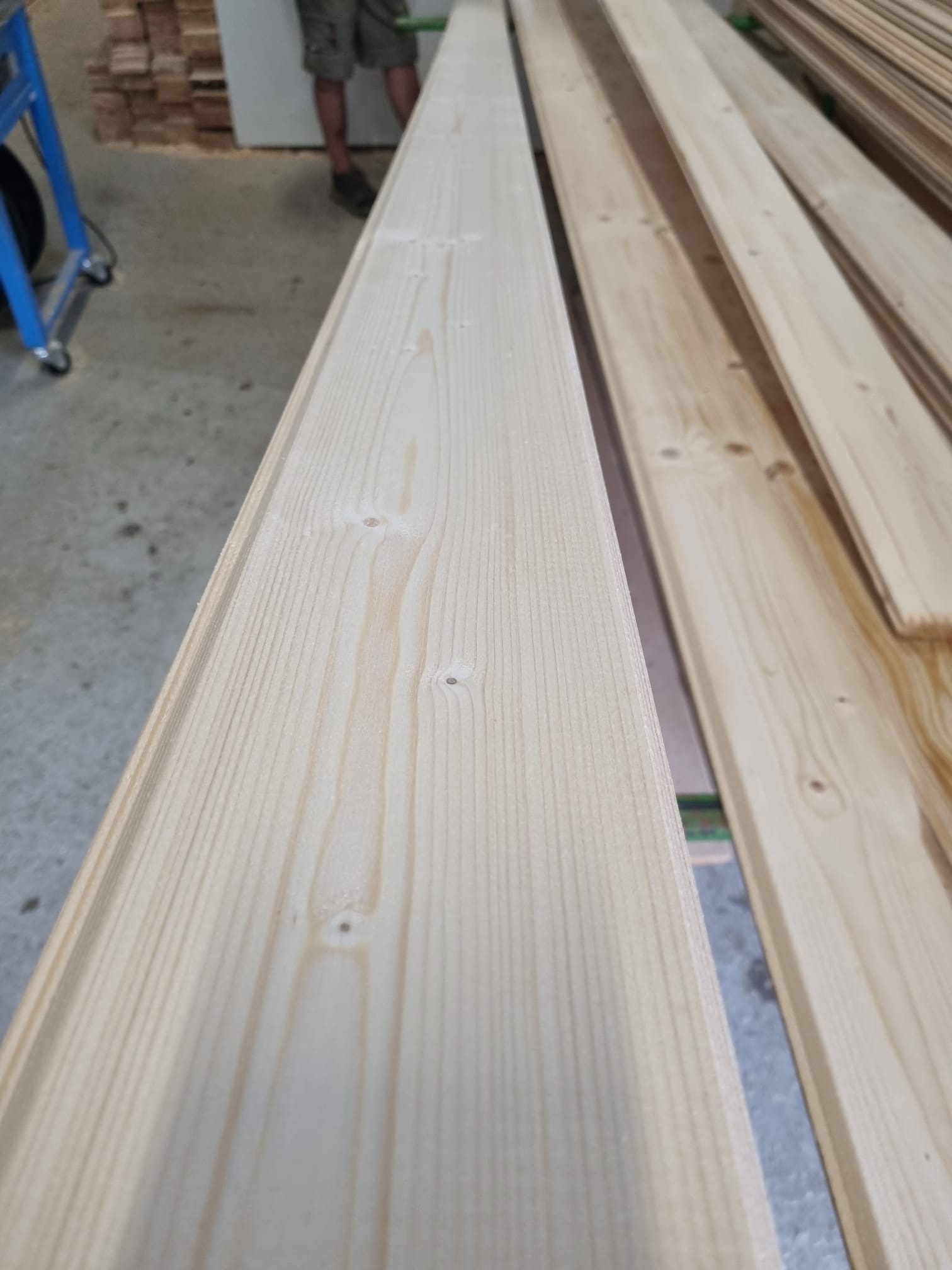 Jasne, heblowane deski z widocznymi słojami drewna, ułożone równolegle w magazynie, widoczne fragmenty wózka transportowego i osoby w tle.