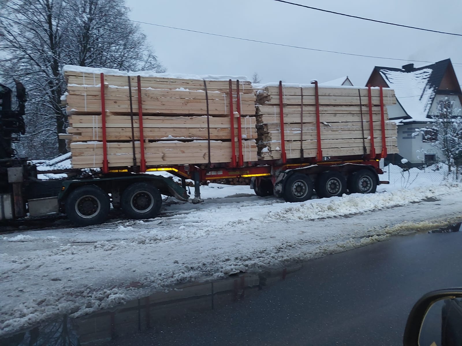 Ciężarówka z przyczepą przewozi ładunek świeżo ściętego drewna budowlanego, zabezpieczonego czerwonymi pasami, w zimowej scenerii.