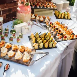 Stół z białym obrusem zastawiony różnorodnymi przekąskami: kanapeczkami z serem, koreczkami z owocami, mini sałatkami w szklankach, ciastkami i tortem w tle.
