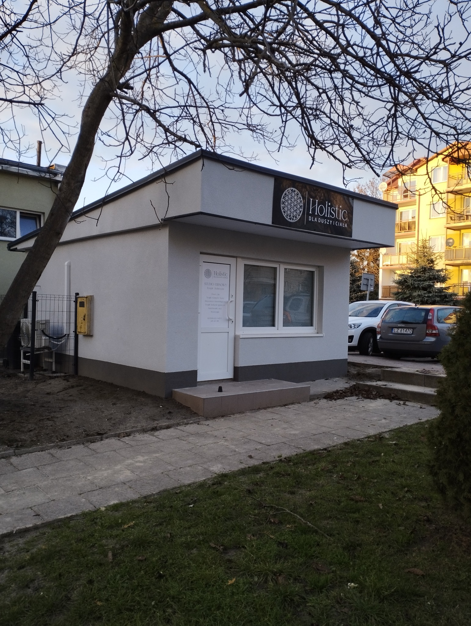 Budynek gabinetu Holistic dla duszy i ciała z logo nad wejściem, widok z zewnątrz, drzewo z nagimi gałęziami na pierwszym planie.