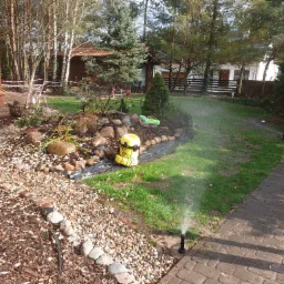 Automatyczny zraszacz trawnikowy w ogrodzie z kamienną rabatą, widoczny żółty pojemnik na nawóz, w tle budynek i drzewa.