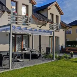 Nowoczesna, biała altana z rozsuwanym zadaszeniem, przylegająca do jasnego domu dwupiętrowego, z widocznym kompletem mebli ogrodowych z rattanu i zadbanym trawnikiem.