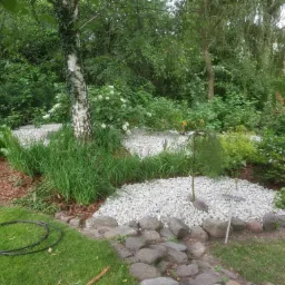 Aranżacja ogrodu z białym grysem, obrzeżami z kamieni polnych i korą, widoczny wąż ogrodowy na trawniku.