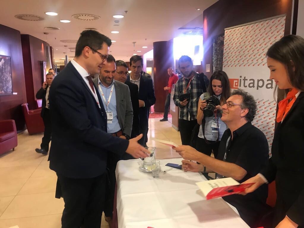 Autor książki podpisuje egzemplarze podczas spotkania autorskiego, otoczony przez osoby z obsługi i fotoreporterów.