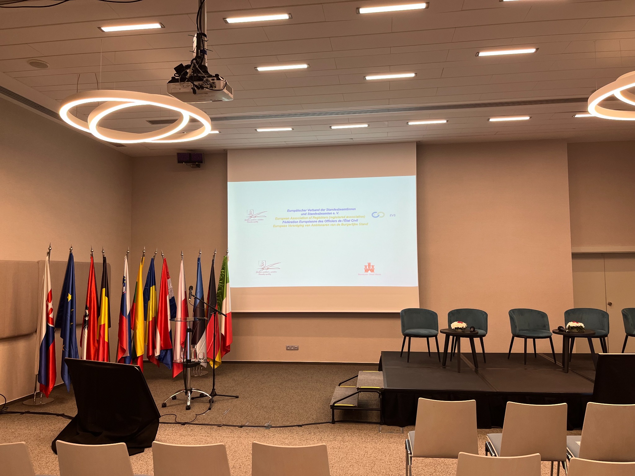 Sala konferencyjna z rzędem flag państw europejskich po lewej stronie, projektor i ekran z tekstem w tle, krzesła dla publiczności na pierwszym planie.