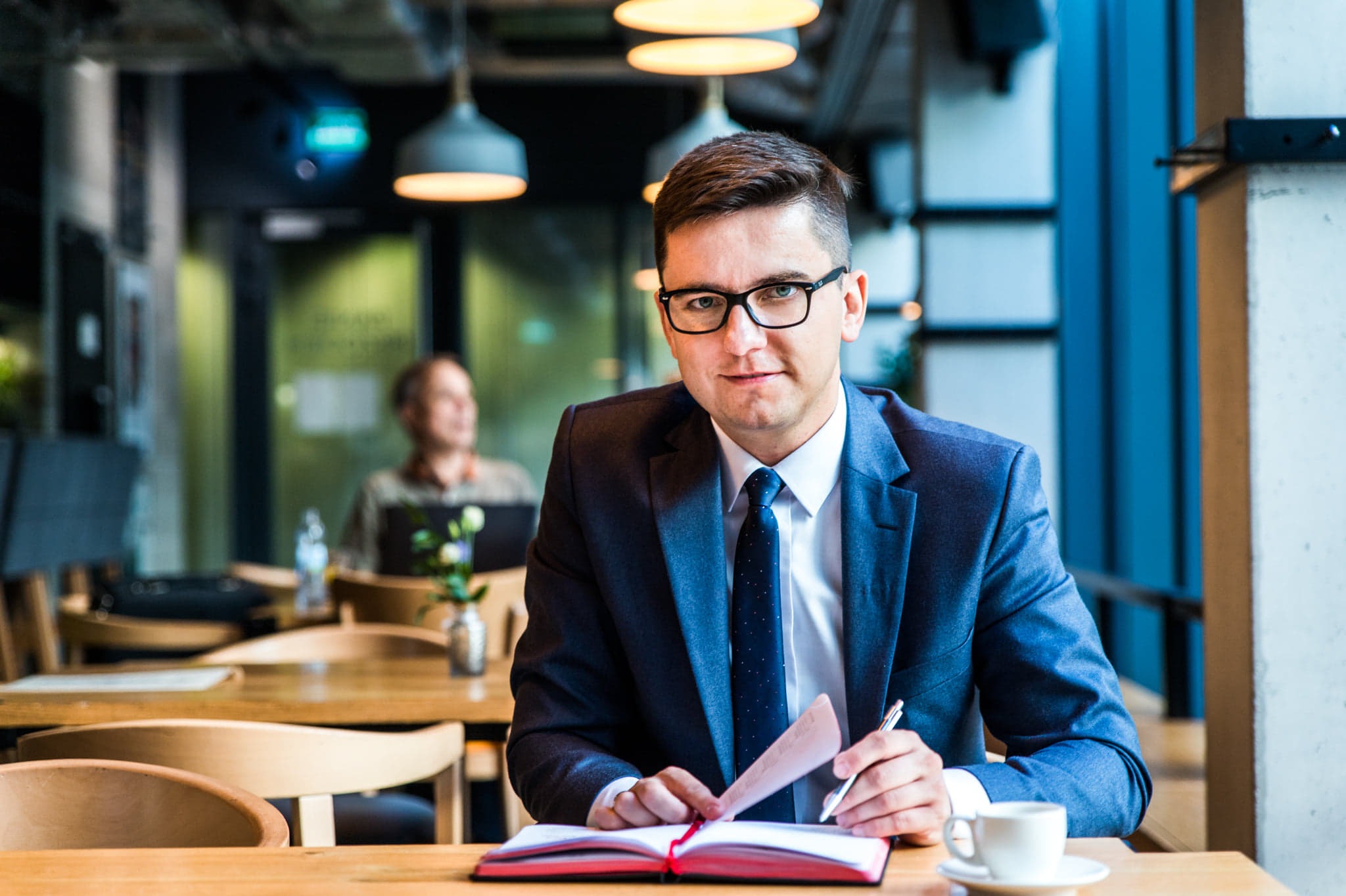 Mężczyzna w garniturze, z okularami, siedzi przy stole w kawiarni w Lublinie, przegląda dokumenty i robi notatki w czerwonym notatniku, obok filiżanka kawy.