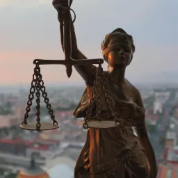 Statua Temidy, bogini sprawiedliwości z wagą, na tle panoramy miasta w ciepłym świetle zachodzącego słońca. Ujęcie od dołu, podkreślające symbolikę.