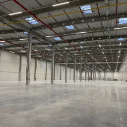 Wnętrze pustej hali magazynowej z betonową posadzką, widoczne kolumny wspierające strop, system rur pod sufitem i szereg lamp oświetleniowych.